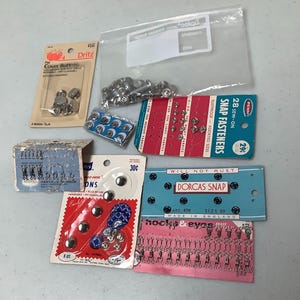 May include: A collection of vintage sewing notions including Dritz cover buttons, Penn sew-on snap fasteners, Scovill hooks and eyes, Dorcas snaps, and loose metal fasteners in a plastic bag. These items are displayed on their original cardboard packaging.