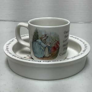 May include: White ceramic mug and saucer set featuring illustrations of Peter Rabbit characters. The mug has a handle and the saucer has a raised rim with text. The set is likely for children or collectors.