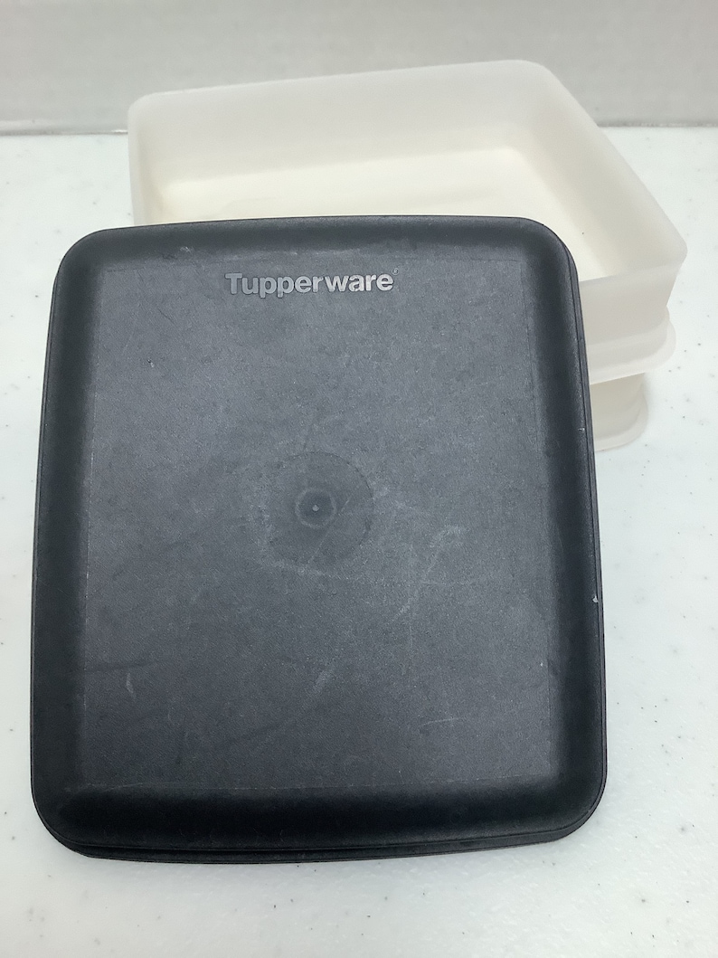 Puede incluir: Un recipiente Tupperware negro con forma cuadrada y esquinas redondeadas. El logotipo de Tupperware es visible en la tapa. Un recipiente blanco rectangular est&aacute; en el fondo.