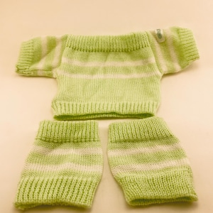 May include: A knitted green and white striped outfit, including a short-sleeved top and two matching leg warmers. The set is made of a soft, textured yarn, with horizontal stripes of white and light green. The outfit is designed for a baby or small child.
