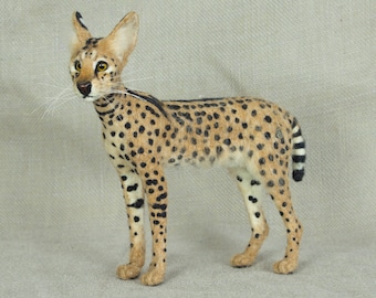 May include: A needle felted serval cat figurine. The figurine is standing on a beige fabric background. The cat is brown with black spots and a long, black-tipped tail.
