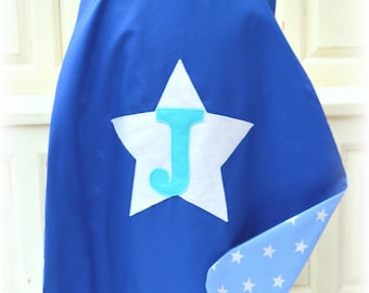 May include: A royal blue cape featuring a large white star appliqué on the back with a light blue letter J centered on the star. The interior lining of the cape is light blue with a repeating pattern of small white stars throughout the fabric.