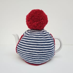 Peut inclure: Une théière en céramique blanche avec un chauffe-thé rayé bleu et blanc. Le chauffe-thé a un pompon rouge sur le dessus et une bordure rouge. La théière est sur une surface blanche.