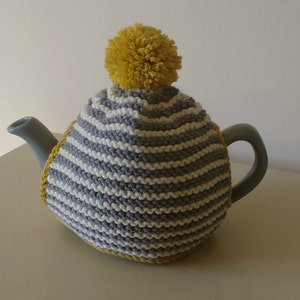 Pode incluir: Um aquecedor de chaleira de malha listrada cinza e branca com um pompom amarelo no topo.