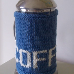Puede incluir: Una cafetera de émbolo de acero inoxidable con una funda de punto azul. La funda tiene la palabra "COFFEE" tejida en letras blancas. La cafetera tiene tapa y base plateadas.