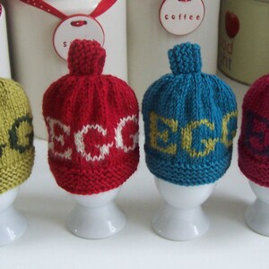 May include: Four knitted egg cozies in different colors, each with the word "EGG" knitted in a contrasting color. The cozies are displayed on white egg cups.