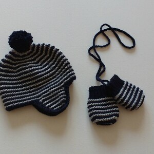 May include: A knitted navy blue and gray striped hat with ear flaps and a matching pom-pom, paired with a set of navy blue and gray striped mittens connected by a navy blue cord. The set is designed for a baby or toddler.