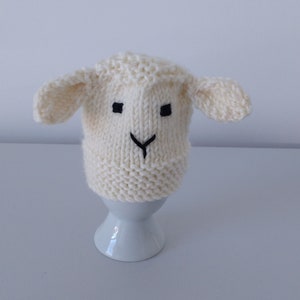 May include: A white knitted lamb hat with black embroidered eyes and nose. The hat is sitting on a white egg cup.