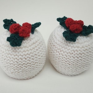 May include: Two white knitted ornaments, each with a sprig of green leaves and three red berries. The rounded ornaments have a knitted texture, ideal for festive decorations. Each measures approximately 8 cm in diameter.