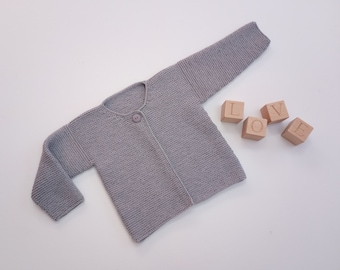 May include: A light gray knitted baby cardigan with a single button closure. The cardigan is made of a soft, textured yarn. Wooden blocks spell out the word "LOVE" next to the cardigan.
