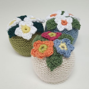 Peut inclure: Trois ornements de pot de fleurs tricotés avec des fleurs de différentes couleurs. Le premier pot est vert avec des fleurs blanches, le deuxième est bleu avec des fleurs blanches et le troisième est blanc avec des fleurs orange, roses et bleues.