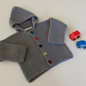 Könnte beinhalten: Grauer gestrickter Kapuzenpullover mit bunten Knöpfen. Der Pullover hat eine Kapuze und einen Reißverschluss vorne.