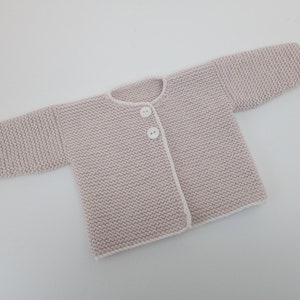 Pode incluir: Um casaco de tricô rosa claro para bebê com botões brancos e uma borda branca ao redor das bordas.