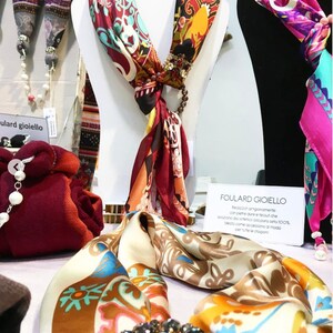 Op de afbeelding: Zijden sjaals met ingewikkelde patronen in rood, oranje en blauwgroen worden op paspoppen getoond. E&eacute;n sjaal heeft een juwelen broche en een kralen accent. De tekst "FOULARD GIOIELLO" is zichtbaar.