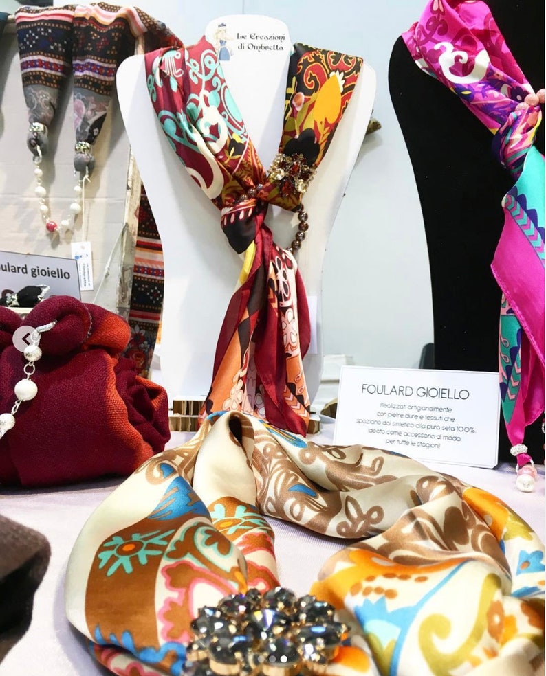 Op de afbeelding: Zijden sjaals met ingewikkelde patronen in rood, oranje en blauwgroen worden op paspoppen getoond. E&eacute;n sjaal heeft een juwelen broche en een kralen accent. De tekst "FOULARD GIOIELLO" is zichtbaar.