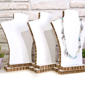 Op de afbeelding: Drie witte sieraden display bustes met een kartonnen basis. E&eacute;n buste toont een lange ketting met zilveren bedels en blauwe en witte kralen. De achtergrond is een witte bakstenen muur met kleine potplanten.