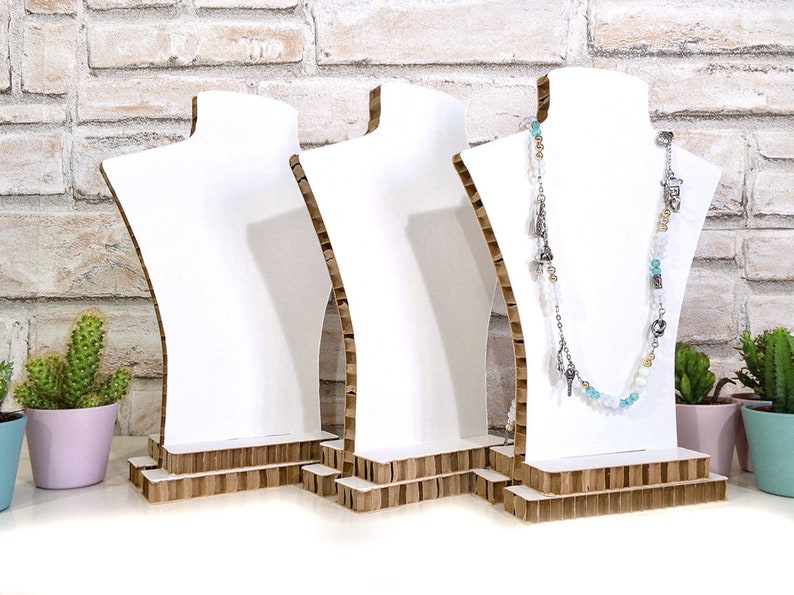Op de afbeelding: Drie witte sieraden display bustes met een kartonnen basis. E&eacute;n buste toont een lange ketting met zilveren bedels en blauwe en witte kralen. De achtergrond is een witte bakstenen muur met kleine potplanten.