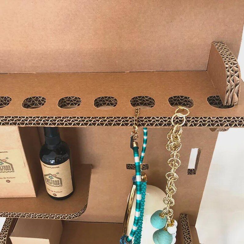 May include: A brown cardboard display unit with a perfume bottle, jewelry, and a box. The unit has oval cutouts and a shelf. The jewelry includes a gold chain and a turquoise beaded necklace. The perfume bottle has a brown label.