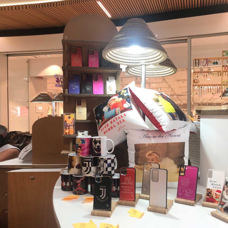 May include: A display of various gift items, including phone cases, mugs, and pillows. The phone cases come in various colors and designs, some with the Juventus logo. The mugs feature different images, and the pillows have printed designs and text. A lamp with a woven shade is also visible.
