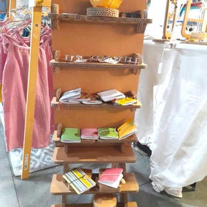 May include: A display stand made of corrugated cardboard, showcasing various notebooks and small items on its shelves. The stand is brown and has multiple levels. Some notebooks have colorful covers. Other items are visible on the shelves.