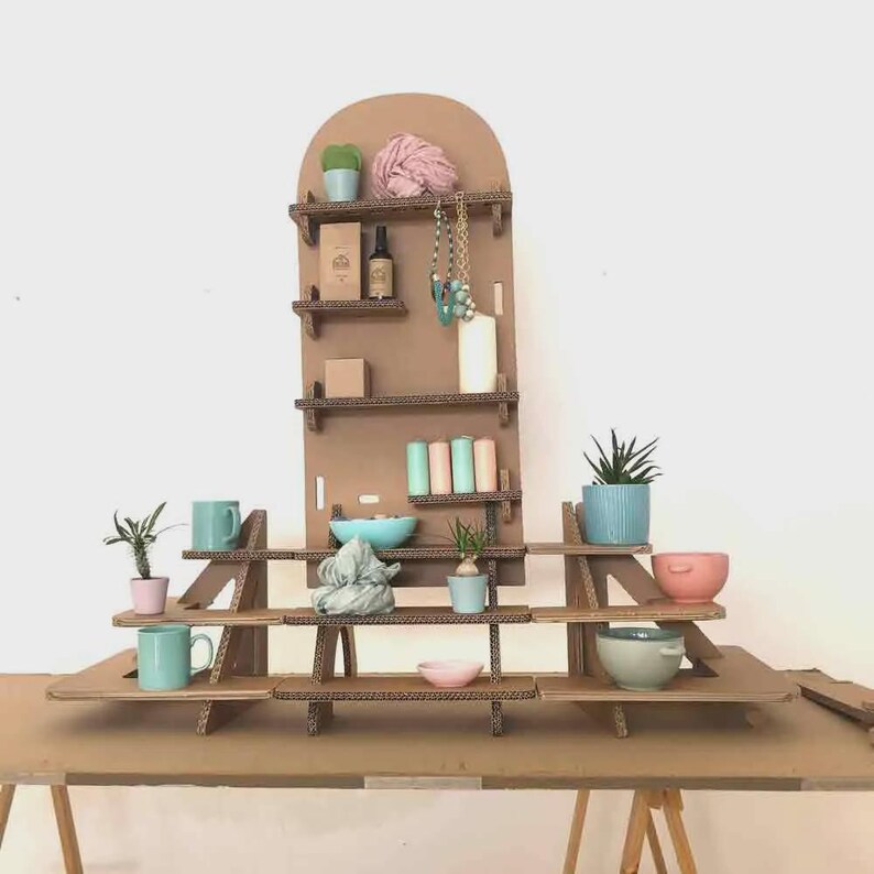 May include: A multi-tiered display shelf constructed from corrugated cardboard. The shelves hold various items, including small potted plants, mugs, bowls, and decorative objects. The display is set against a plain white background.