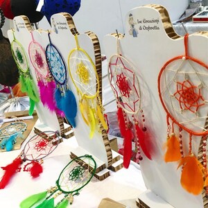 Op de afbeelding: Een display van kleurrijke dromenvangers met ingewikkelde webontwerpen en levendige veren. De dromenvangers zijn in verschillende kleuren, waaronder groen, roze, blauw, geel en rood. De tekst "Ise Creazioni di Ombretta" is zichtbaar.
