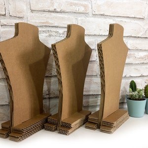 Op de afbeelding: Drie bruine kartonnen kettingdisplay stands. De stands zijn gevormd als een buste met een basis. De achtergrond is een witte bakstenen muur. Twee kleine cactussen in potten in lichtblauwe potten staan rechts.