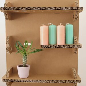 May include: A three-tiered, corrugated cardboard shelf unit. The shelves hold four pillar candles in pastel pink and teal, and a small potted plant. The unit is made of brown cardboard with decorative edges.