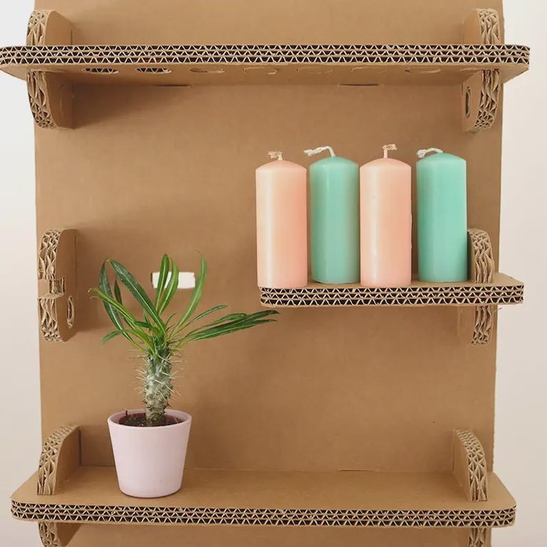 May include: A three-tiered, corrugated cardboard shelf unit. The shelves hold four pillar candles in pastel pink and teal, and a small potted plant. The unit is made of brown cardboard with decorative edges.