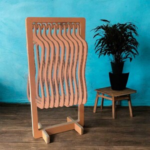 May include: A brown cardboard clothing rack with a series of wavy hanger slots. The rack is freestanding with a rectangular base and a tall, rectangular top. A potted plant sits on a small wooden table in the background.