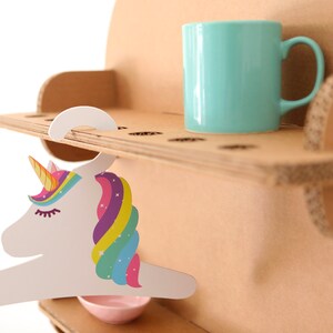 May include: A cardboard shelf unit with a light blue mug and a unicorn-shaped hanger. The unicorn hanger has a colorful mane and horn. A small pink bowl is below the hanger. The shelf unit is made of corrugated cardboard.