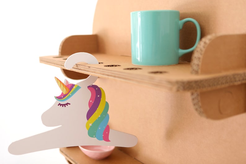 May include: A cardboard shelf unit with a light blue mug and a unicorn-shaped hanger. The unicorn hanger has a colorful mane and horn. A small pink bowl is below the hanger. The shelf unit is made of corrugated cardboard.