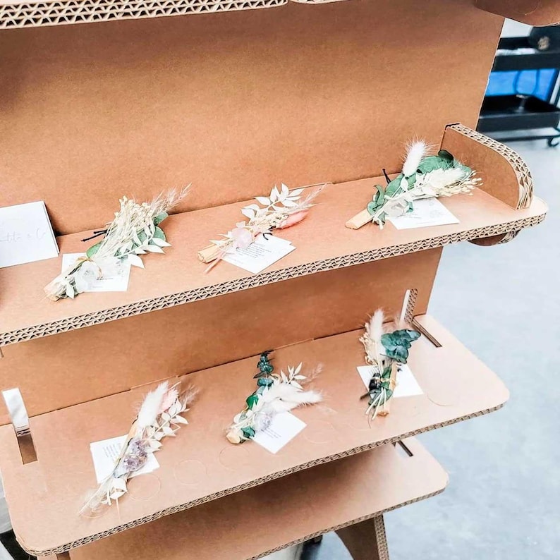 May include: A display of dried flower bouquets arranged on cardboard shelves. The bouquets feature a variety of textures and colors, including white, green, and pink. Each bouquet is accompanied by a small white card. The display is set up in a retail environment.