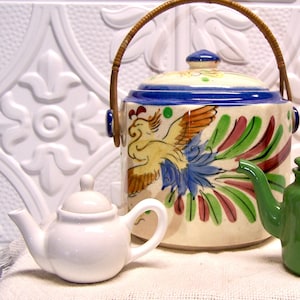 May include: A white ceramic teapot with a lid and a handle. The teapot is sitting on a white surface with a larger, colorful ceramic teapot with a bird design in the background.