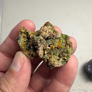 May include: A close-up of a mineral specimen featuring a cluster of small, yellow, crystal formations. The mineral has a rough, textured surface with green and brown hues. The specimen is held in a hand.