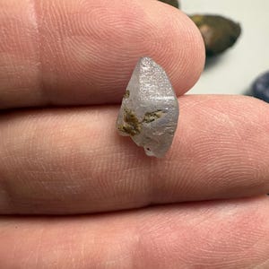 May include: A small, light purple crystal with a pointed tip and rough texture. The crystal has some brown inclusions and is held between fingers. The background is blurred, with other small stones visible.
