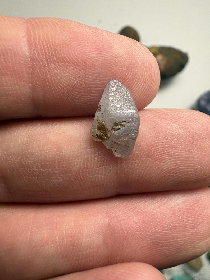 May include: A small, light purple crystal with a pointed tip and rough texture. The crystal has some brown inclusions and is held between fingers. The background is blurred, with other small stones visible.
