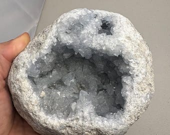 May include: A large, rough-textured, light grey geode with a hollow centre filled with pale blue crystal formations. The geode is held in a hand, showcasing its size and natural beauty. The crystals sparkle, contrasting with the stone's exterior.