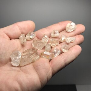 May include: A collection of small, clear quartz crystals with a slightly golden hue. The crystals are faceted and have a rough, natural texture. They are displayed in a hand, showcasing their size and shape.