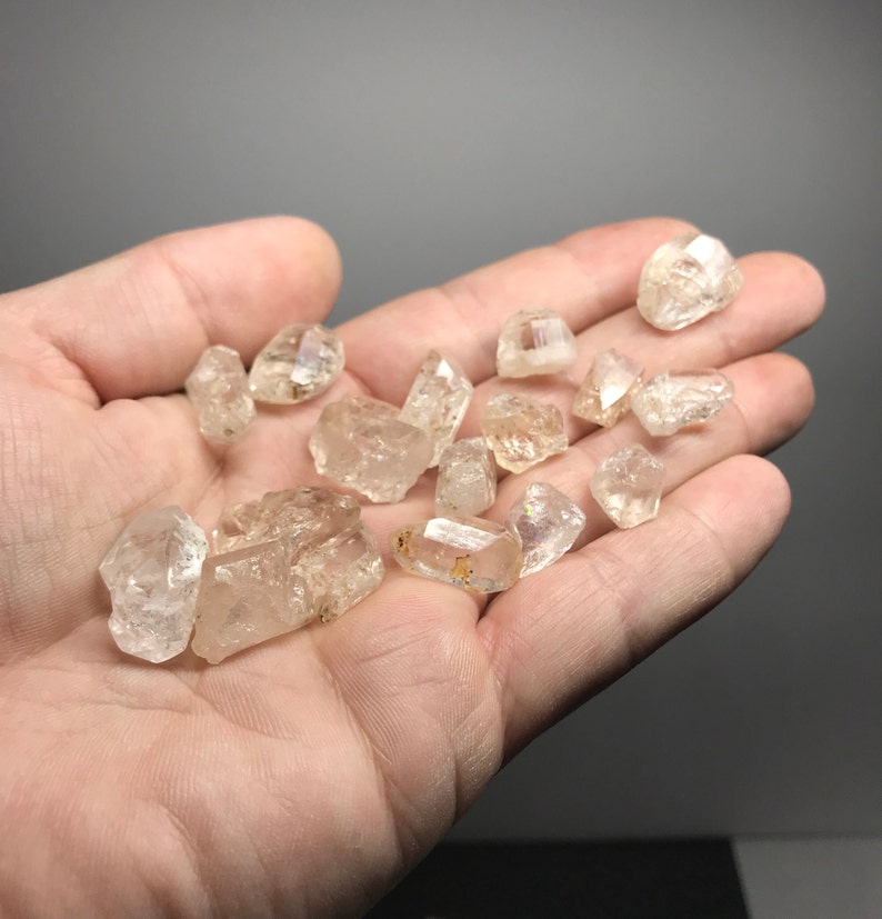 May include: A collection of small, clear quartz crystals with a slightly golden hue. The crystals are faceted and have a rough, natural texture. They are displayed in a hand, showcasing their size and shape.