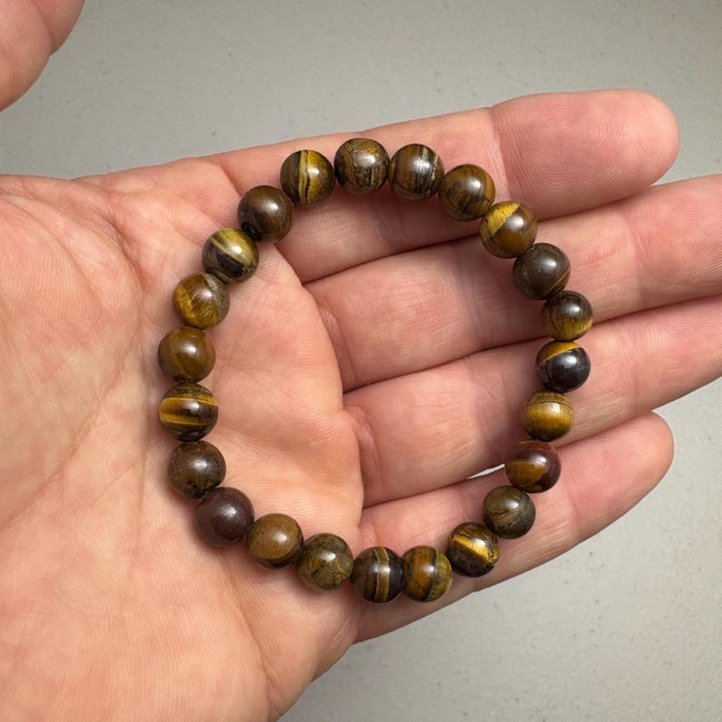 May include: A bracelet made of round, polished tiger eye beads. The beads are primarily brown with bands of yellow and gold. The bracelet is elastic and designed to be worn on the wrist.