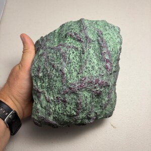 May include: A large, rough-textured rock with a green base colour and deep red veins running throughout. The rock is held in a hand, showcasing its size and natural patterns. This geological specimen is a unique decorative item.