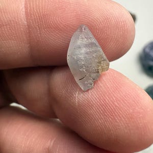 May include: A small, translucent, light blue crystal with a pointed top and a rough, textured surface. The crystal has a slightly irregular shape and appears to be a natural gemstone. The crystal is held between fingers.
