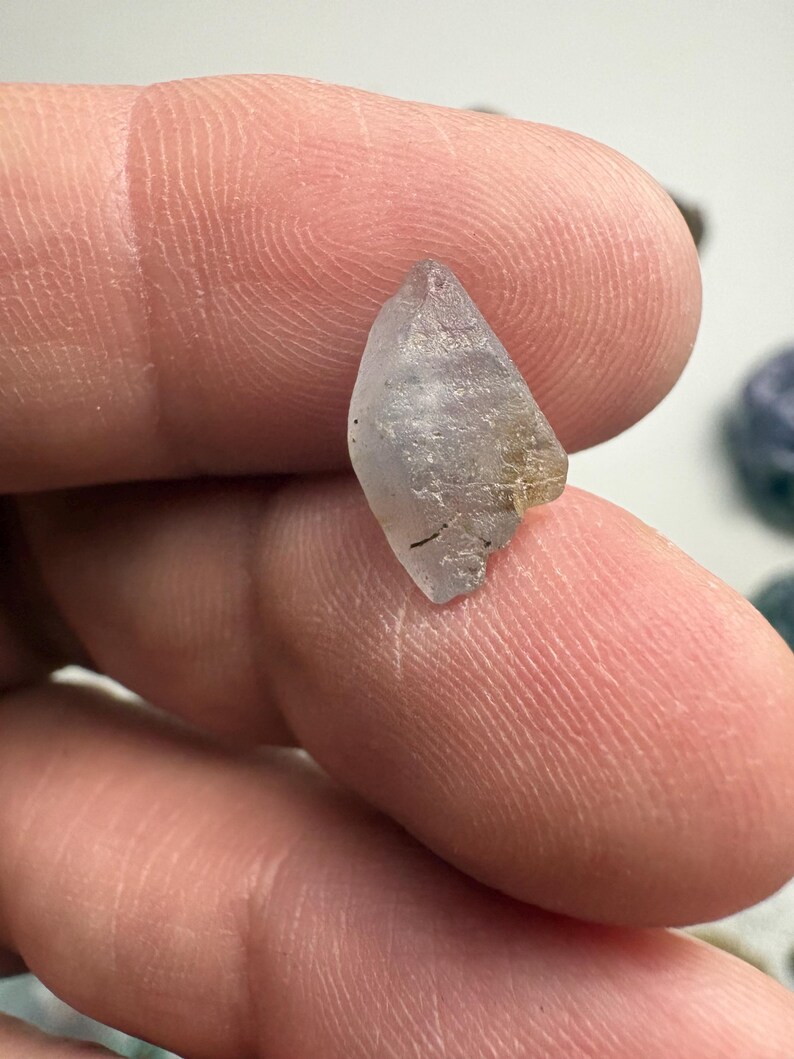 May include: A small, translucent, light blue crystal with a pointed top and a rough, textured surface. The crystal has a slightly irregular shape and appears to be a natural gemstone. The crystal is held between fingers.