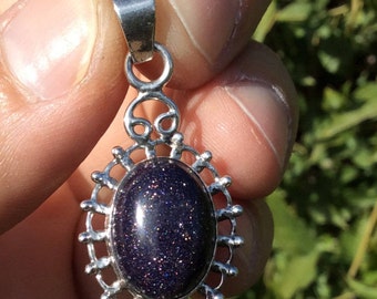 May include: A silver pendant with an oval, dark blue stone speckled with glitter. The stone is set in a decorative silver frame with an ornate bail. The pendant is held up against a green, leafy background.