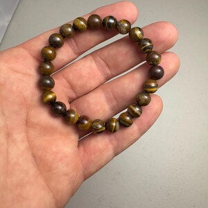 May include: A beaded bracelet made of tiger's eye stones. The bracelet features round, polished beads in shades of brown, gold, and black. The bracelet is strung on an elastic band, and is a stretch bracelet. The bracelet is shown on a hand.
