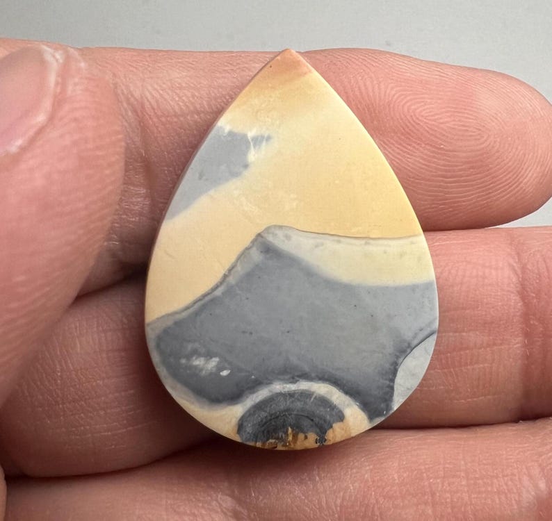 May include: A teardrop-shaped cabochon of polished stone with a swirling pattern of gray, white, and yellow.