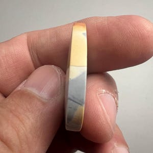 May include: A close-up of a round, multi-colored stone cabochon with a yellow, gray, and white pattern. The stone is being held by a hand.