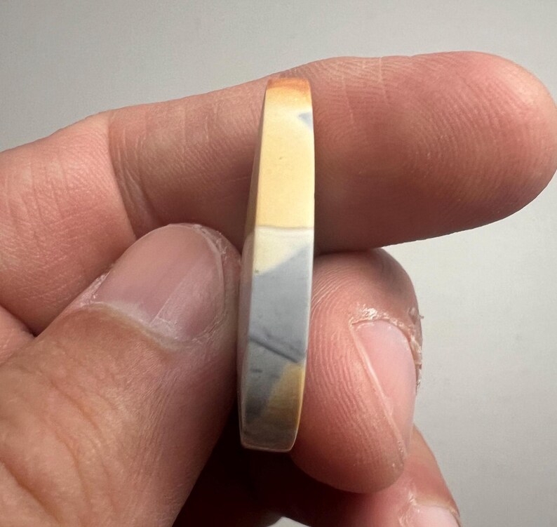May include: A close-up of a round, multi-colored stone cabochon with a yellow, gray, and white pattern. The stone is being held by a hand.