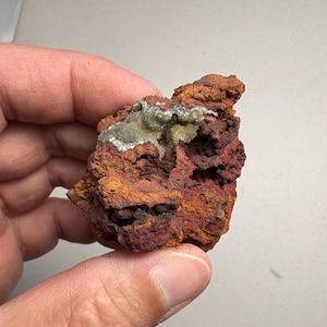 May include: A rough, reddish-brown rock formation with areas of orange and dark red. The rock has a porous texture and a patch of light green and white mineral deposits.